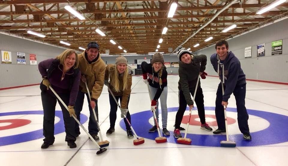 default-101 Two men and four women with sweeping brooms stand on the painted circles of a curling rink.