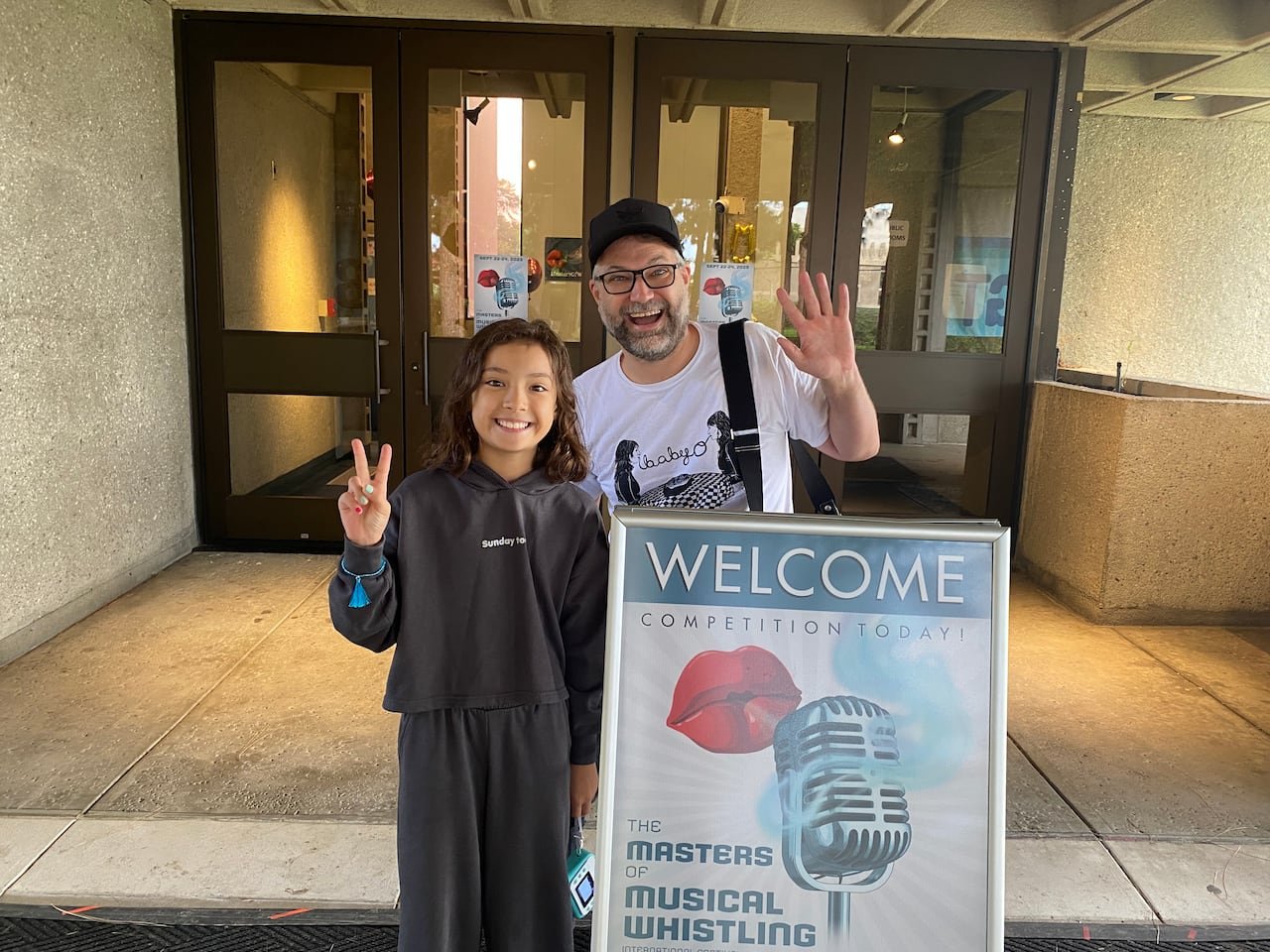 default-288 An adult man and young girl wave in front of a stone building's entrance. They're standing behind a sandwich board sign with the word "WELCOME" on it and a painting of an old fashioned microphone.