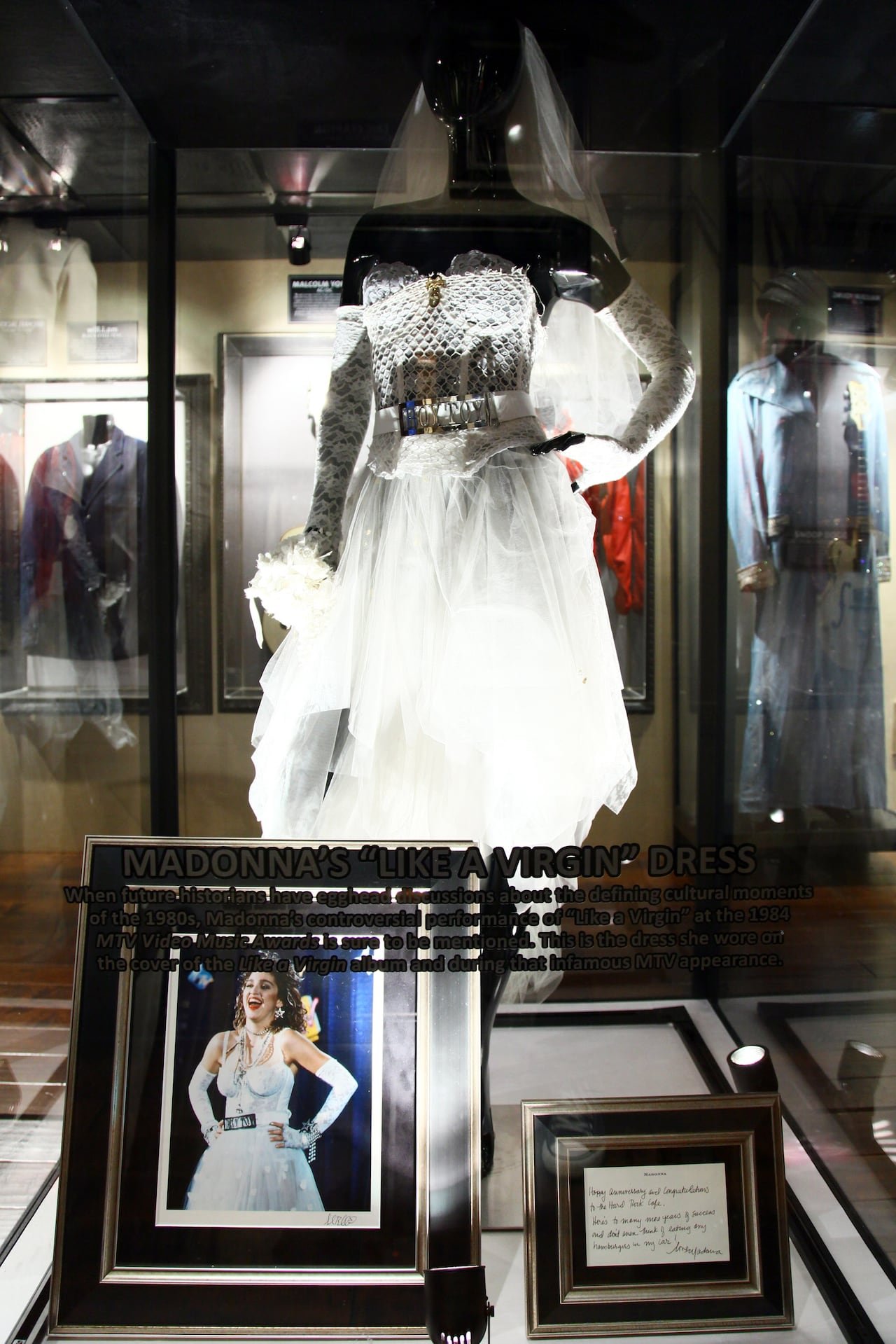 default-229 A wedding dress and a belt reading "Boy Toy" displayed in a case on a mannequin. A photo of a woman wearing the dress and a plaque sit in front of it.